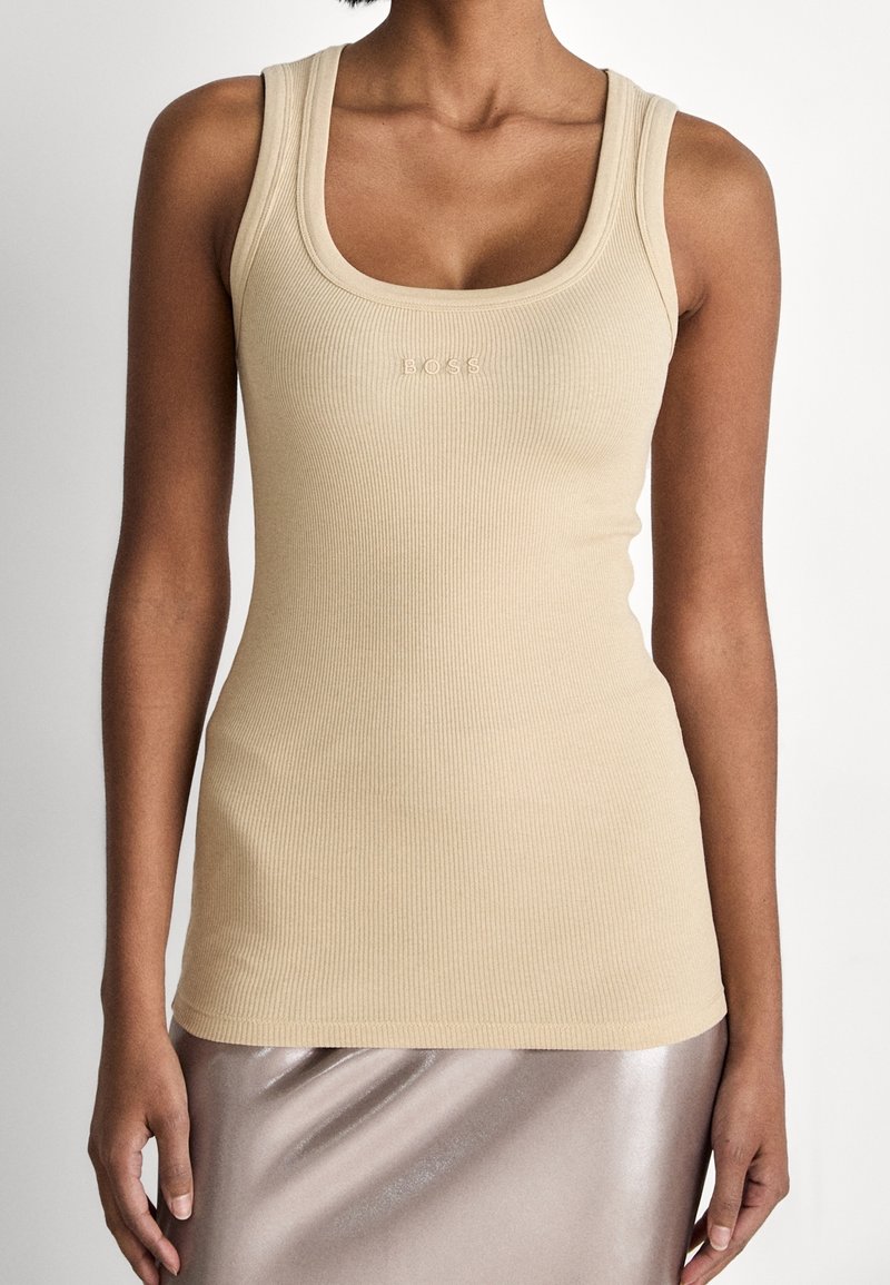 Kvinna som bär en beige ribbad linne med "BOSS"-logotyp präglad på bröstet och en glänsande ljus taupe-kjol.