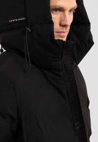 Giacca a vento nera con un grande cappuccio. Presenta una finitura testurizzata, zipper frontale e logo embossato lungo la patta. Isolamento leggero.