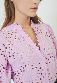 Light pink blouse featuring intricate cut-out patterns, scalloped collar and button closure, with textured fabric and a fitted design.