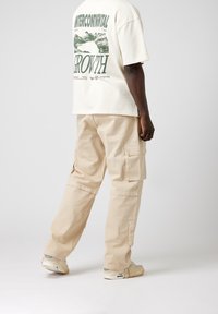 Beige cargobyxor med sidofickor, kombinerade med en krämfärgad t-shirt med grafik som har grön text och berg. Lättviktig bomullstyg.