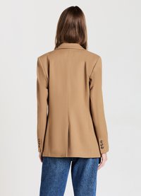 Blazer beige su misura con rever a punta, spacco singolo sul retro e polsini abbottonati, abbinato a jeans blu a vita alta.