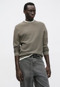 Grå strikket sweater med ribbet struktur, rund halsudskæring og lange ærmer, båret over en hvid skjorte. Kombineret med grå bukser.