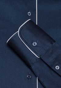Navy blue shirt with white accents, featuring a button-down collar, button closures, and textured fabric. Cuff folded over the sleeve.