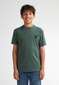 Groen katoenen T-shirt met korte mouwen, ronde hals en een klein zwart vierkant patch op de linkerborst. Casual pasvorm, gladde textuur.