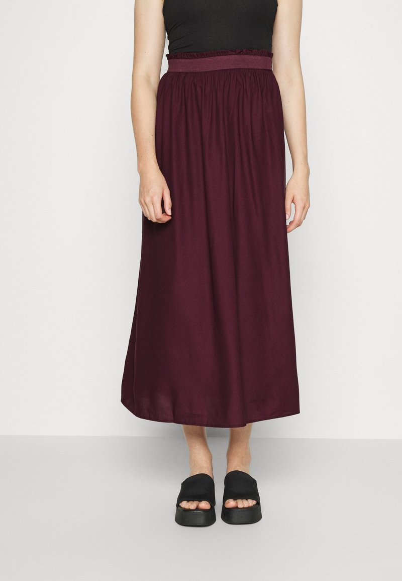 Femme portant une jupe prune à taille haute, froncée, longueur cheville, avec un haut sans manches noir et des sandales à plateforme noires.