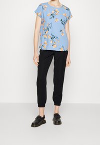 En person står klädd i en blå blommig topp med orange blommor och svarta smala byxor, kompletterad med svarta skor, mot en neutral bakgrund.