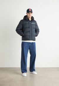Giacca a vento nera con cappuccio, chiusura con bottoni e texture liscia, abbinata a jeans blu larghi e sneakers bianche. Gli accessori includono un cappellino.