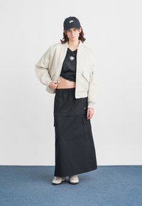 Witte bomberjack, zwarte mesh top, zwarte lange rok met zakken en een zwarte pet. Casual sneakers maken de outfit compleet.