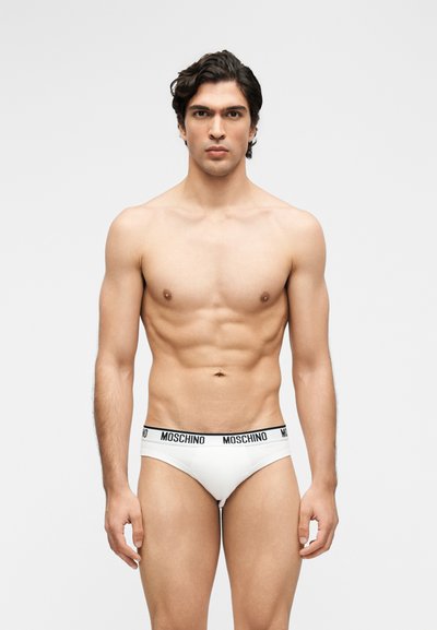 Μυώδης άνδρας με κοντά σκουρόχρωμα μαλλιά, όρθιος, φοράει λευκά εσώρουχα Moschino με μαύρη λωρίδα μέσης με λογότυπο σε μονόχρωμο φόντο.