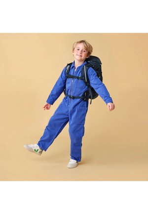 Kleines Kind im blauen Overall mit schwarzem Rucksack, lächelnd, hebt ein Bein, steht vor beigem Hintergrund.