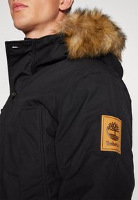 Parka noire avec une capuche en fourrure, dotée d'un écusson en cuir Timberland et d'une fermeture à boutons-pression. Tissu texturé et poches latérales.