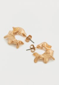 Σκουλαρίκια κρίκοι σε χρυσό τόνο με υφή, με οργανικό, κυματιστό σχέδιο και κούμπωμα με καρφίδα. Αναδεικνύουν ακανόνιστες μορφές και γυαλιστερές επιφάνειες.