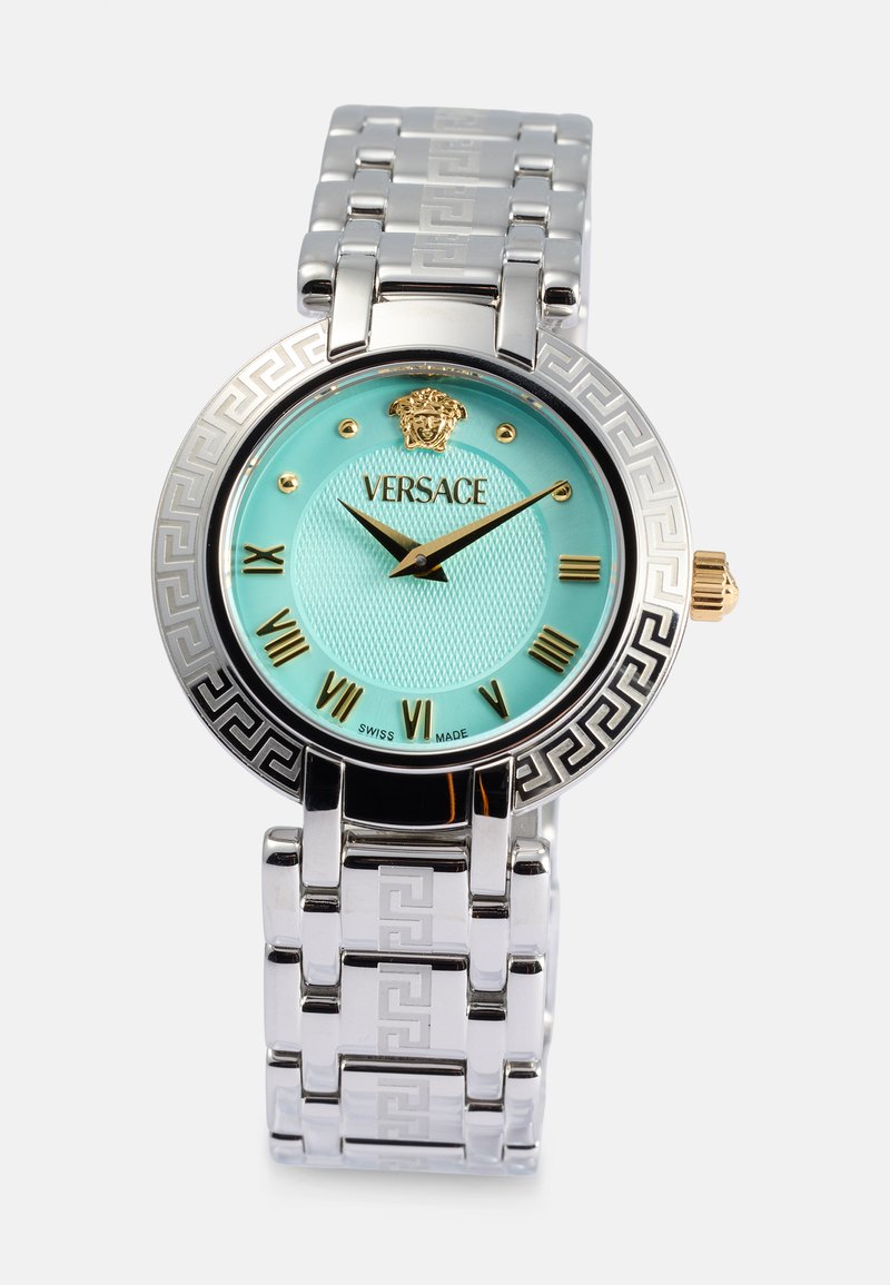 Orologio in metallo argento con quadrante testurizzato color acquamarina, dettagli dorati, indicatori con numeri romani e un motivo a chiave greca sulla lunetta. Marchio Versace incluso.
