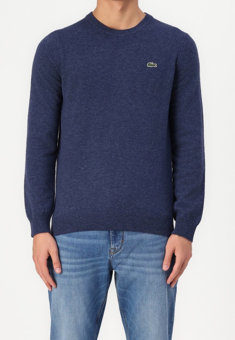 Mörkblå stickad tröja med långa ärmar, rund halsringning och subtil struktur. Har en liten grön krokodil-logotyp på bröstet.