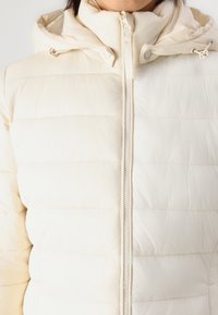 Chaqueta acolchada en color crema con cierre frontal, costuras horizontales y capucha. El material parece suave y ligeramente brillante.