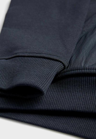 Materijal tamnoplave boje s rebrastom teksturom; sadrži preklopni manžet i blago sjajnu tkaninu, ističući slojevite detalje tkanine.