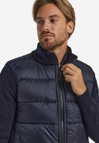 Granatowa pikowana kamizelka z wysokim kołnierzem, zamkiem z przodu oraz rękawami w stylu swetra z prążkowanego materiału. Materiał jest gładki, z pikowanym wypełnieniem.