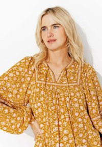 Blouse à manches longues, jaune moutarde, avec un motif floral en rose et blanc. Elle présente un décolleté en V et des coutures décoratives le long du empiècement.