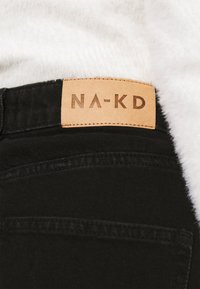 Schwarze Denim-Jeans mit einem braunen Lederetikett, das den eingeprägten Text "NA-KD" am Bund trägt. Die Textur ist glatt und strapazierfähig.
