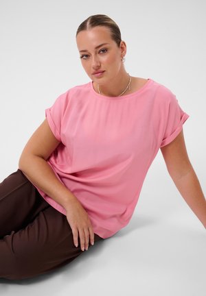 Femme aux cheveux plaqués en arrière portant une chemise rose à manches courtes et un pantalon marron, assise par terre devant un fond blanc uni.