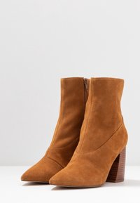 Botas de tobillo de ante marrón con punta afilada y tacón apilado, cuentan con una cremallera lateral. La textura es suave, con líneas limpias y modernas.
