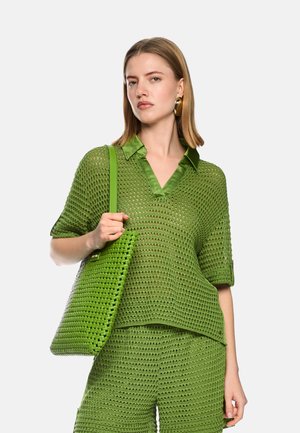 Woman wearing green knit top and shorts, holding matching woven green tote bag over shoulder against plain background.