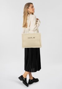 Beige shearling totebag met een reliëf "REPLAY"-logo, voorzien van twee bovenhandvatten en een zachte, getextureerde oppervlakte. Het model draagt een zwarte geplooide rok.