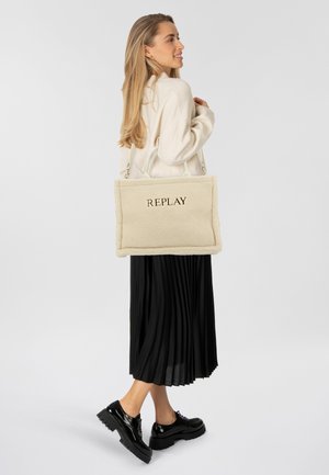 Beiger Shearling-Shopper mit geprägtem "REPLAY"-Logo, zwei oberen Griffen und einer weichen, strukturierten Oberfläche. Das Modell trägt einen schwarzen Plisseerock.
