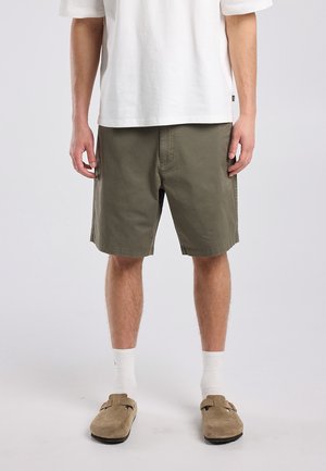 Man wearing olive green shorts, a white t-shirt, white crew socks, and beige slip-on shoes, standing against a plain light background.