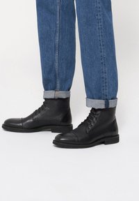 Czarne skórzane buty wiązane z okrągłym noskiem, teksturowanym wierzchem i płaską podeszwą, w zestawie z niebieskimi dżinsami z podwiniętymi mankietami.