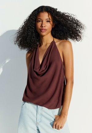 Jeune femme aux cheveux bouclés portant un haut marron sans manches à col halter et un jean en denim bleu clair, devant un fond uni clair.