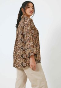 Brown floral-patterned oversized shirt with three-quarter sleeves and lace accents, paired with light beige pants. Smooth fabric texture.