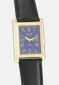 Montre rectangulaire Casio avec un boîtier en acier inoxydable doré, un cadran bleu marine, des chiffres romains et un bracelet en cuir noir. Résistante à l'eau jusqu'à 50 mètres.