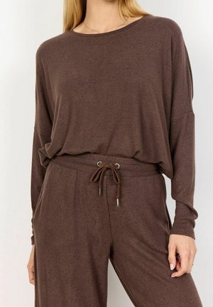 Mujer con una blusa marrón de manga larga suelta metida en pantalones a juego con cordón y bolsillos, de pie frente a un fondo liso.