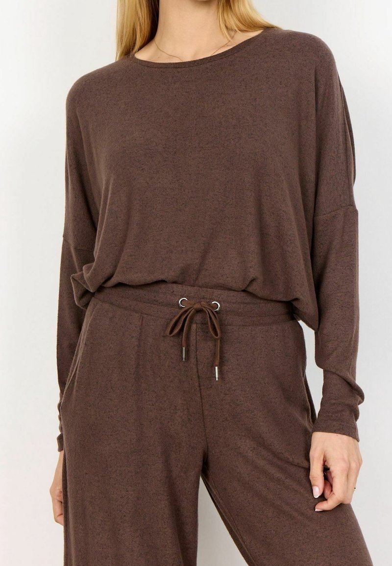 Femme portant un haut marron ample à manches longues, rentré dans un pantalon à cordon assorti avec des poches, debout devant un fond uni.