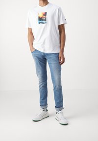 Vit bomullströja med färggrafisk tryck, tillsammans med ljusblå denimjeans och vita sneakers med subtila gröna accenter.