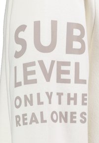 Weißes Sweatshirt mit fettem, hellgrauem Text, der "SUB LEVEL ONLY THE REAL ONES" liest. Sanfter Stoff mit entspannter Passform.