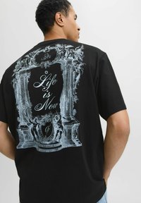 Czarna koszulka z krótkim rękawem z srebrnym nadrukiem przedstawiającym ozdobne kolumny i anioły oraz napisem "Życie jest Teraz" na środku. Luźny krój.