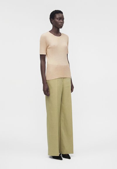 Γυναίκα όρθια, φοράει μπεζ ριπ κοντομάνικη μπλούζα, φαρδιά ανοιχτό ελιά παντελόνια και μαύρα μυτερά παπούτσια σε απλό φόντο.