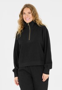 Schwarzer Reißverschluss-Pullover aus strukturiertem Stoff mit gerippten Details und hohem Kragen. Mit einem goldenen Reißverschluss und lockerer Passform.