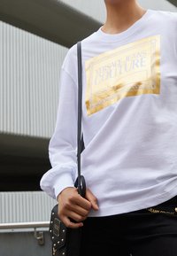 Bílá košile s dlouhým rukávem s barevným grafickým designem a nápisem "VERSACE JEANS COUTURE". Modelka drží černou kabelku s cvočky.