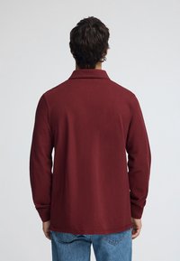 Vinröd långärmad pikétröja med krage, med slät textur och avslappnad passform, kombinerad med blå denimjeans.