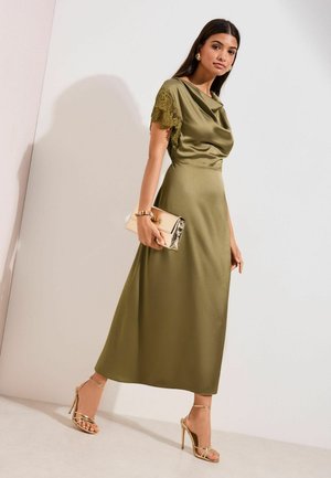 Kvinde iført olivengrøn satin kjole med blonder korte ærmer, holder en gylden clutch og har på gyldne sandaler med høje hæle mod en hvid væg.