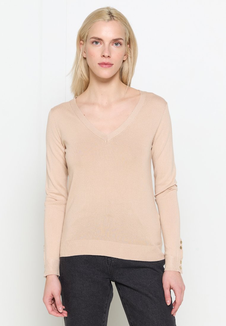 Suéter de punto color beige con cuello en V, mangas largas, textura acanalada y detalles de botones en los puños, combinado con jeans gris oscuro.