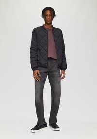 Zwarte gewatteerde bomberjack, donker bordeauxrood t-shirt, grijze jeans. De broek is iets vervaagd. Zwarte sneakers met witte zolen maken de outfit compleet.