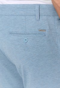 Světlé modré kalhoty z texturované látky. Mají zadní kapsu se zapínáním na knoflík a malou značkovou cedulku u švu kapsy.