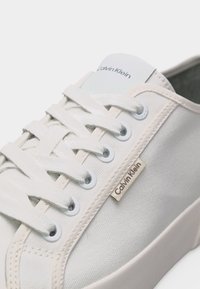 Λευκό αθλητικό παπούτσι Calvin Klein με λευκά κορδόνια, μεταλλικές τρύπες και λογότυπο της μάρκας στο πλάι και στη γλώσσα.