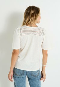Camiseta de manga corta color crema con un panel de encaje texturizado en los hombros. Material suave, corte relajado y escote redondeado.