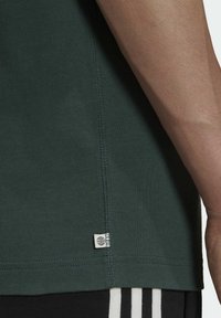 Nahaufnahme des Arms einer Person und dem Saum eines dunkelgrünen Shirts mit einem kleinen weißen Etikett, das ein Globus-Symbol zeigt, kombiniert mit schwarzen Hosen mit weißen Streifen.