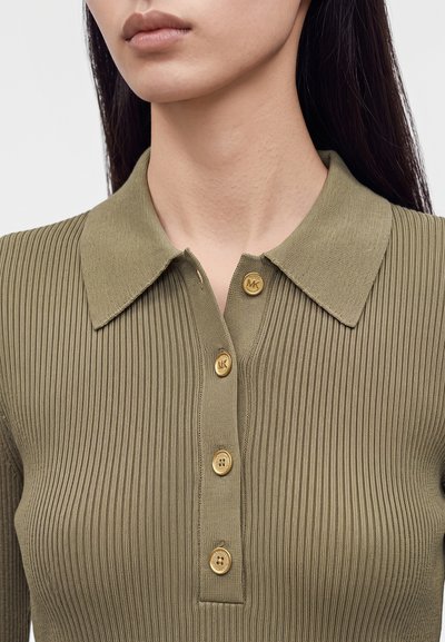 Gros plan d’une personne portant un polo en maille côtelée vert olive avec un col et quatre boutons dorés sur le devant.